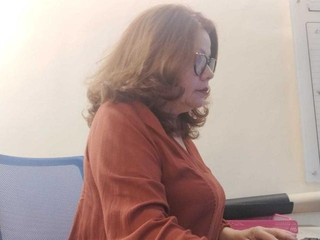 CristelleDeLuxe, middle-aged woman with brown curly hair, wearing glasses and a red cardigan, sitting in an office chair, holding a black pen, looking to the right.