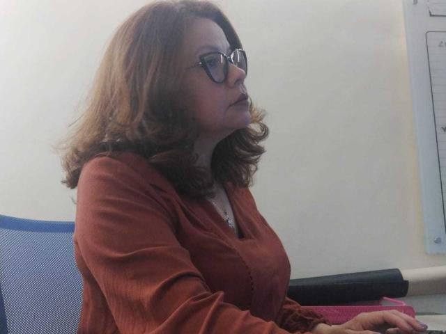 CristelleDeLuxe with wavy brown hair, glasses, and a red long-sleeve shirt, sitting in an office chair, looking to the right, with a white wall and desk behind.