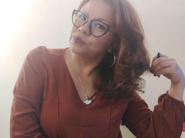 CristelleDeLuxe with wavy auburn hair, wearing black glasses, a red blouse, and a necklace, holding a black pen in her right hand, looking slightly to the left.