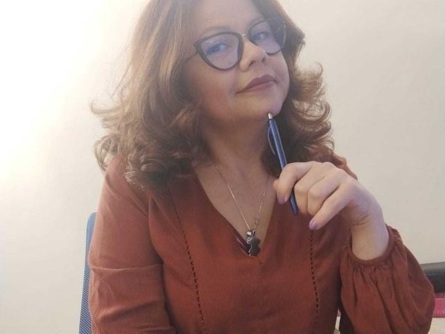CristelleDeLuxe with wavy brown hair, glasses, and red top, holding a blue pen to her chin, slightly smiling. Boobs are not visible, but she's looking cute and smart.