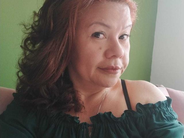 CristelleDeLuxe, a middle-aged woman with curly red hair, green off-shoulder top, and black strap, smiling slightly, showing small tits and a cute mole on her right cheek.