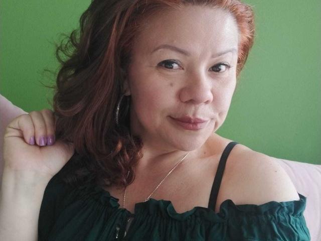 CristelleDeLuxe has red wavy hair, wearing a green off-shoulder top, showing her tits with black straps, purple nails, and a silver necklace, smiling at the camera.