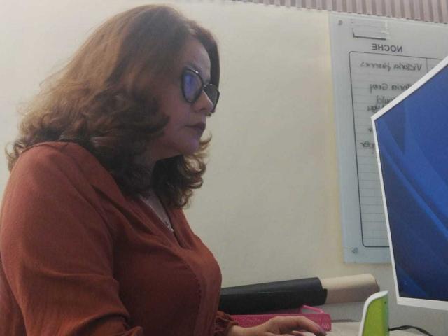 CristelleDeLuxe has curly brown hair, wears glasses, and a red blouse, sitting with her tits slightly visible, looking to the right, next to a computer and green cup.