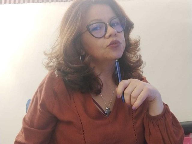 CristelleDeLuxe with curly brown hair, glasses, pouty lips, holding a pen near her mouth, wearing a rust-colored shirt, looking slightly annoyed. Boobs are partially visible under the shirt.