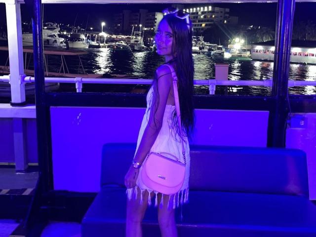 TamaraPeach se tient dans un bateau éclairé par une lumière bleue, vêtue d'une robe blanche à franges, tenant un sac rose, avec de longs cheveux bruns et un sourire discret, et regardant par-dessus son épaule..