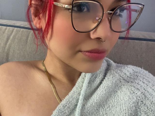 SharonFenix with red hair, glasses, nose ring, and gold necklace, wearing a light blue towel, showing her bare shoulders and a slight pout, looking at the camera.
