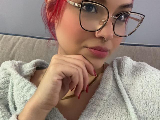 SharonFenix with red hair, glasses, nose ring, and red nails, wearing a fluffy gray robe, resting her chin on her hand, showing off her pink lips.