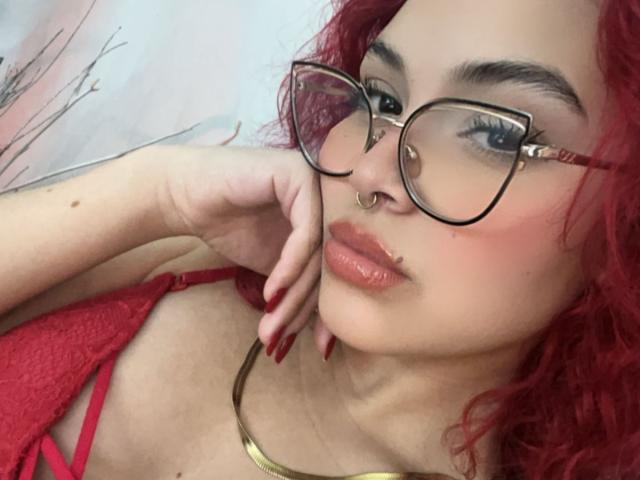 SharonFenix with curly red hair, wearing red lace bra, gold necklace, glasses, nose ring, and lip ring, has red nails, and a slightly flushed face, looking seductive.