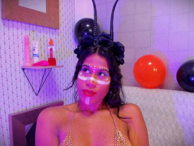 GodessMar, con un copricapo a forma di corno nero, trucco bianco sul viso e top da bikini dorato, ha i capelli scuri, rossetto rosso e si trova in una stanza con palloncini e un sex toy su uno scaffale..