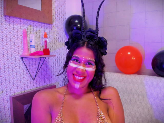 GodessMar with dark curly hair, wearing black horn headpiece, white face paint, and leopard print bikini, smiling with pink and purple lighting, red and black balloons in background.