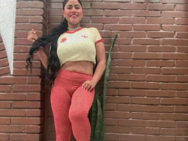 IvannaDuran with long black hair, yellow crop top showing her tits, and red pants, standing against a brick wall, smiling with a hand in her hair.