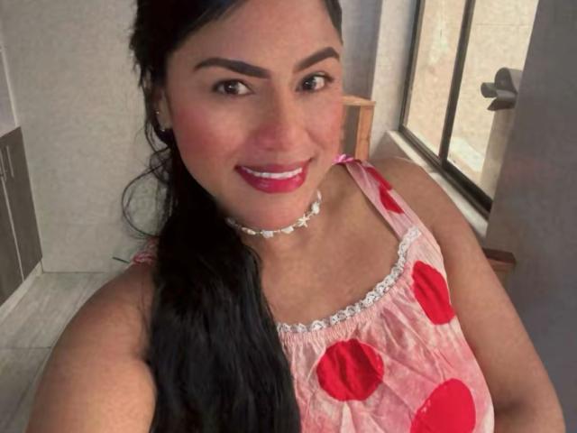 IvannaDuran with long black hair, red lipstick, and a pink dress with red polka dots, wearing a white beaded choker, smiling at the camera. Boobs visible under dress.