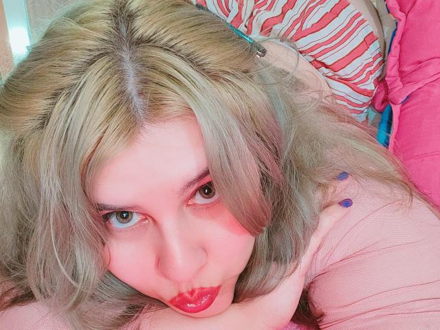 Akkanee has blonde hair, pink skin, red lipstick, and blue nail polish, lying on a pink and striped pillow, looking up with a pouty face.