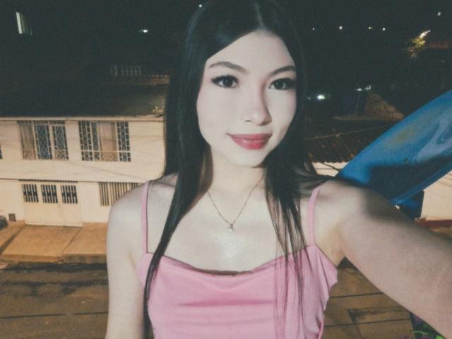 ViolettaLeroy with long black hair, pink tank top, small tits, pale skin, red lipstick, and a subtle smile, standing at night with a blue object behind her.
