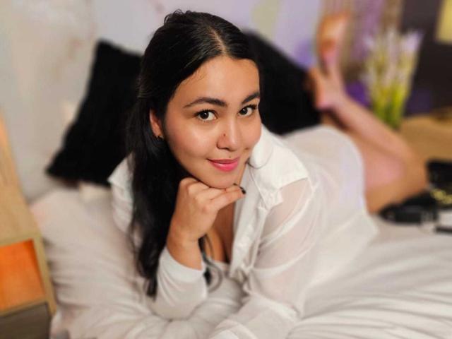 TaiTanakaa with black hair, pink lips, and brown eyes, wearing a white shirt, lying on a bed, smiling, resting her chin on her hand, showing a cute, sexy face.