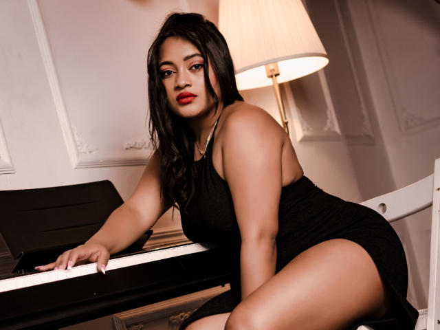 SaritaCute with big ass, black dress, red lipstick, and long black hair, leaning on piano, looking at camera, white chair, lamp in background, sexy and confident.