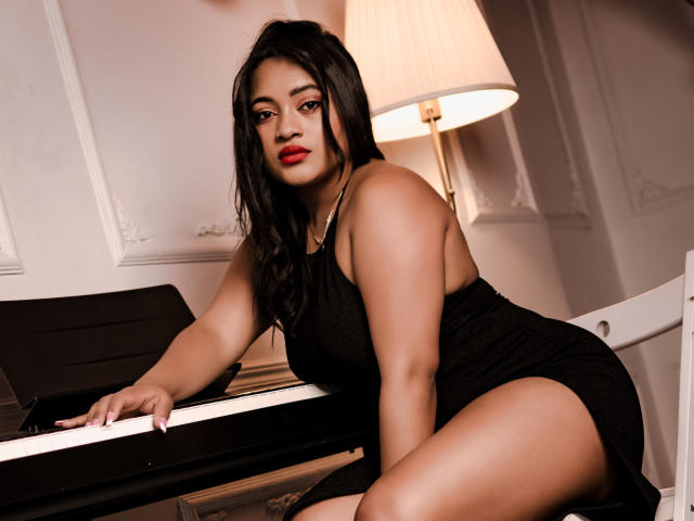 SaritaCute, com seu corpo avantajado e seios bonitos, está apoiada em um piano, vestindo um vestido preto, com batom vermelho e cabelos longos e pretos, ao lado de um abajur em um quarto branco..