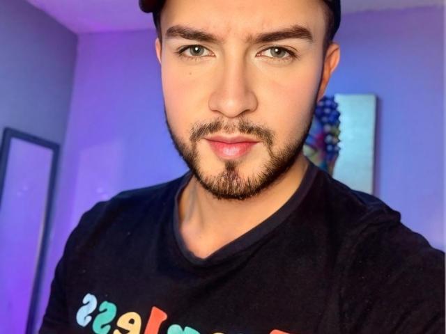 DaltonWhite, a cute dude with a short beard and mustache, green eyes, and a black cap, wearing a black t-shirt, showing off his smooth, slightly pink, soft-looking ass.