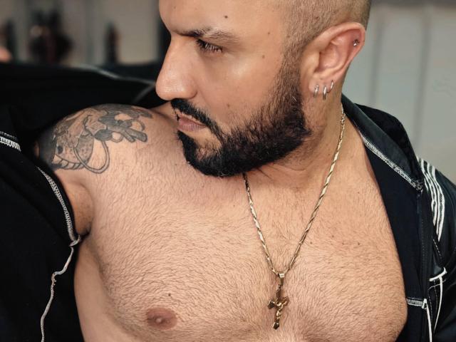 BardoCkroy69 with a shaved head, beard, tattoo on shoulder, chain necklace, and ear piercings, showing hairy chest, black jacket, and looking downward.