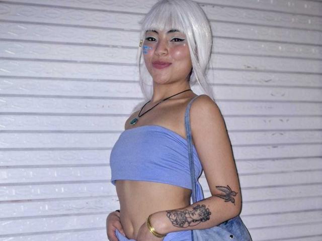 SammyHills69 with white hair, blue tube top, small tits, tattoo on right arm, blue face paint, necklace, and blue purse, standing against white shutter background.
