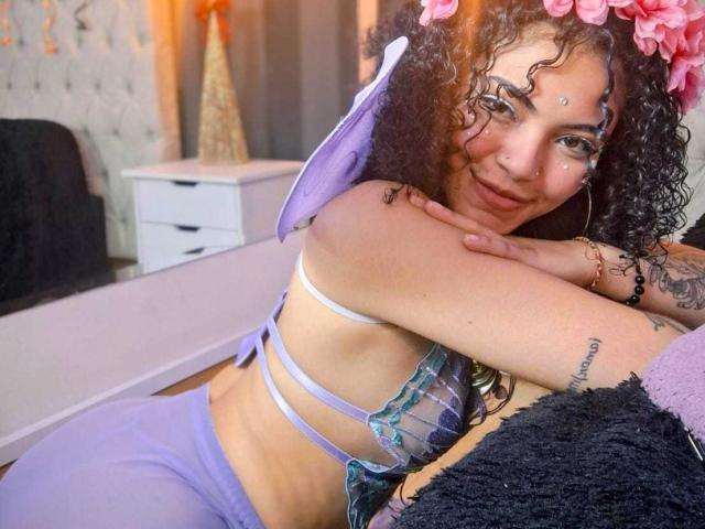 SammyHills69 with curly hair, wearing a purple lace bra and matching pants, leaning on a black pillow, smiling, flower crown, tattoo on right arm, small beauty mark on forehead.