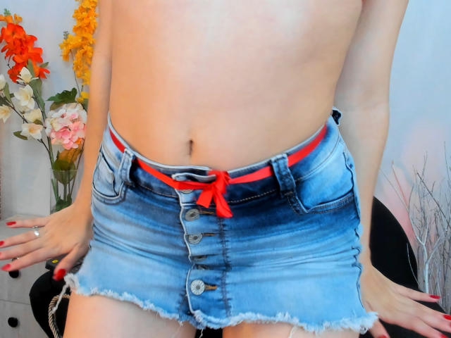SharonFenix's bare midriff with a blue, button-down, frayed denim skirt, red waistband, and red nails. Her hands rest on her thighs, showing a ring.