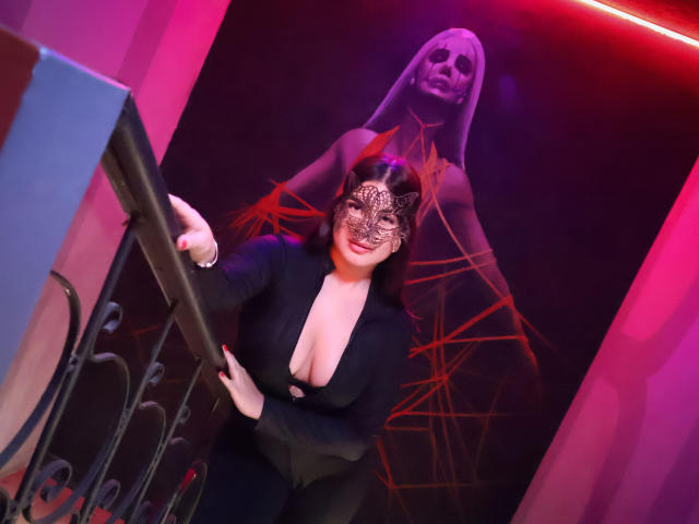 CamilaSofiia with red mask, black dress, and deep cleavage, leaning on a railing, standing in front of a large, red-lit, black-and-white portrait of another masked woman.