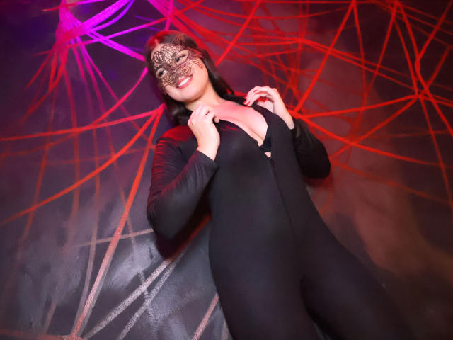 CamilaSofiia in a black, tight, zip-up bodysuit, hands near her chest, wearing a decorative mask, smiling, with red and purple spiderweb lights in the background.
