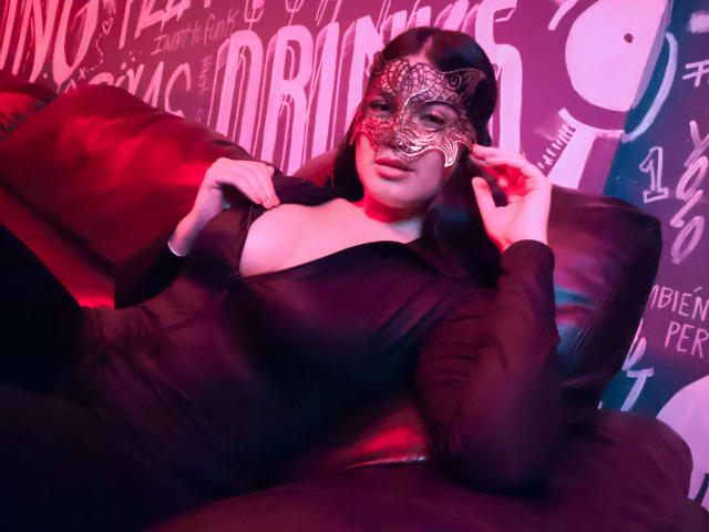 CamilaSofiia with a black mask, black dress, and red lighting, showing off her tits, lying on a black leather couch, hands near her face, looking sexy.