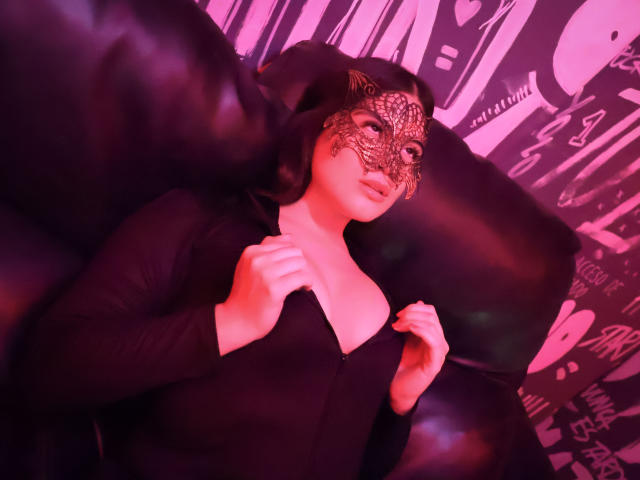CamilaSofiia with black mask, lying on black leather couch, wearing black zip-up dress, hands near chest, pink and purple lighting, short black hair, pale skin, soft expression.