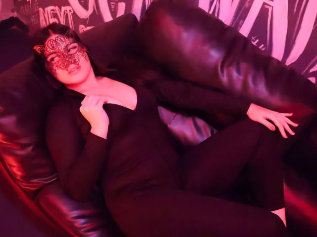 CamilaSofiia in a black bodysuit, lying on a black leather couch, wearing a leopard print mask, with her hand on her chest, and her legs crossed.