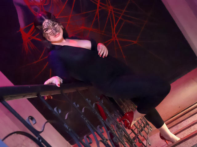 CamilaSofiia in a black dress, mask, and red heels, leaning on a metal railing, with her hand on her belly, and her leg slightly raised.