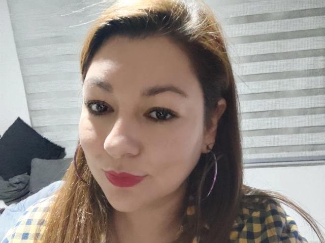 SakotoDika tiene la piel de color marrón claro, lápiz labial rojo y ojos marrones. Lleva una camisa a cuadros amarillos y azules con aros de aro rosas, y está sonriendo ligeramente. Su cabello marrón es lacio..