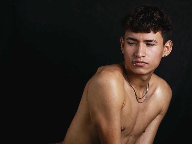 NicolasTorres is a young man with medium brown skin and curly dark hair. He's shirtless, showing a lean, muscular upper body. His right arm is crossed over his chest, and his left arm is slightly raised. He's wearing a simple black necklace and a small earring in his left ear. His expression is neutral, with slightly parted lips. The lighting is soft, highlighting his smooth skin and subtle muscle definition. The background is completely black, making NicolasTorres the focal point. The composition is tight, with NicolasTorres positioned slightly to the right.