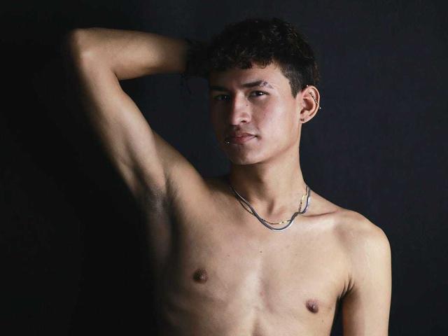 NicolasTorres is shirtless with a chain necklace, arm raised, short curly dark hair, light brown skin, small nipples, and a serious expression, against a dark background.