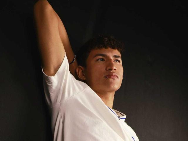 NicolasTorres, a young man with curly brown hair, is leaning against a dark wall. He's wearing a white shirt with a blue trim on the collar. His right arm is raised, and his left arm is partially visible. His facial expression is neutral, with a slight pout. He has a small piercing on his lower lip. The light source is from the front, highlighting his facial features and the texture of his shirt. The dark background contrasts with his light shirt and skin tone. The composition is simple and focused on NicolasTorres' upper body and face.