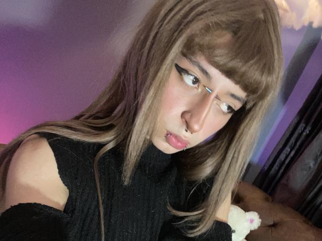 OasisFairy with long brown hair, thick eyeliner, nose ring, and pink lips, wearing a black off-shoulder turtleneck, looking down with a slightly pouty expression.