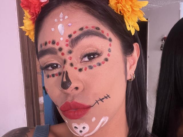 StellaBeckker has dark hair, yellow and red flower headpiece, face painted with skull, cross, and dots, red lipstick, black nose, white and black paint, and a small white bear.