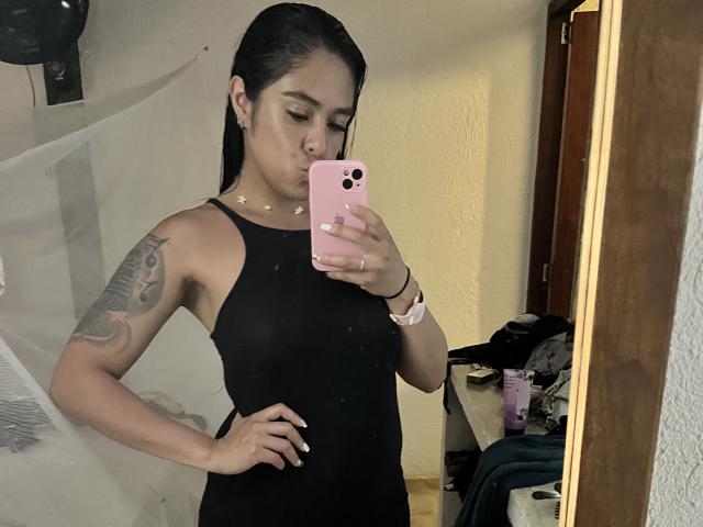 StellaBeckker, with black hair, wearing a black tank top, showing a tattoo on her arm, taking a mirror selfie with her pink phone, pouty face, and hand on hip.