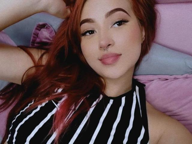 SamaraCuttie, com cabelo ruivo, blusa listrada preta e branca, piercing no nariz e delineador, deitada em almofadas rosa, sorrindo, exibindo seu rosto fofo e nariz pequeno..