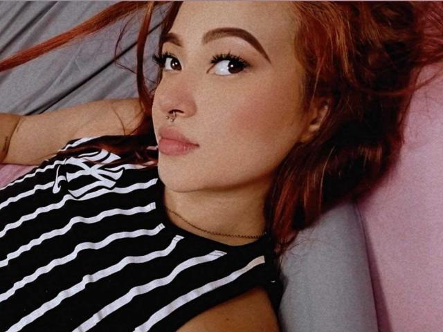Close-up of SamaraCuttie with red hair, lying down. She has a nose ring in her right nostril, thick eyebrows, and long eyelashes. She wears a black and white striped sleeveless top. Her skin is fair with a slight pinkish tint. She gazes slightly to the right with a neutral expression. Her hair is messy, and she's on a gray pillow with a pink pillow behind her. The lighting is soft, highlighting her facial features. The image has a casual, everyday feel.