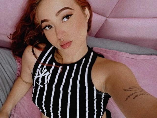 SamaraCuttie has light skin and red hair, lying on a pink bed. She wears a black and white striped sports top with a Nike logo. Her right arm has a tattoo with cursive text. She has a nose ring in her right nostril and dark, winged eyeliner. The composition is a close-up, slightly angled from above. Her expression is neutral, and she looks directly at the camera. The texture of the bed is plush and quilted.