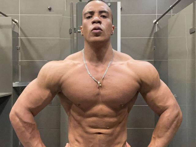 Muscular FrankDickerson with a bald head, wearing a necklace, stands shirtless in a bathroom, showing off a defined chest and large biceps, with a serious expression.