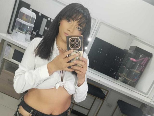 AnnaMooree mit mittelgroßer Brust, in einem weißen Hemd, das zur Hälfte geöffnet ist, schwarze Hose, hält ein Handy, Selfiemit Spiegel, braunes Haar, Perlenkette, leichtes Lächeln, weißer Raum, Spiegelung, Handyhülle mit goldener Verzierung..