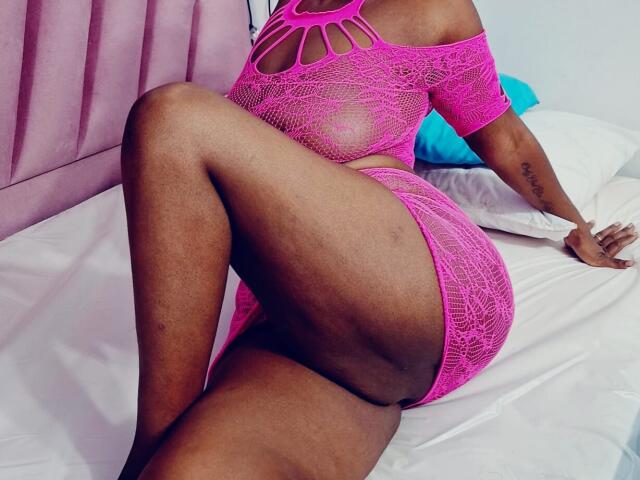 MissFoster in pink lace lingerie, showing her curvy ass and thighs, lying on a bed with her hand on white sheets, her tattoo visible on her right arm.