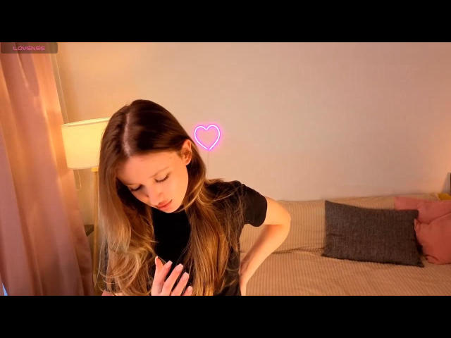 EiraKilian, avec de longs cheveux blonds, un t-shirt noir et un panneau lumineux en forme de cœur rose, penchée en avant, les yeux baissés, la main près de la poitrine, sur un lit avec des oreillers gris et des rideaux roses..