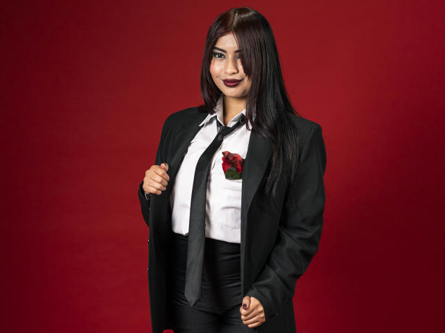 EmmaaRodriguez with long black hair, wearing a black suit, white shirt, black tie, and red rose pin, standing against a red background, smiling with dark lipstick.