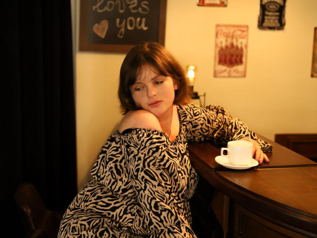 StaceyLex, a fair-skinned woman with short brown hair, leans on a wooden table, holding a white coffee cup on a saucer. She wears a black and white zebra-pattern dress, with one shoulder slightly down. Her expression is neutral, eyes half-closed. The table has a dark wooden texture, and the warm, yellowish light adds a cozy feel. The background includes a chalkboard with "coffee you" in white chalk and other wall decorations. Her right arm rests on the table, while her left hand is out of frame. The image is focused on StaceyLex.