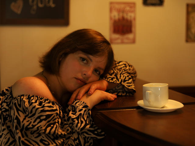StaceyLex leans on a wooden table with her head resting on her hands, wearing a zebra-patterned off-shoulder top. She has short brown hair and a neutral expression. A white ceramic cup and saucer sit on the table to her right. Warm, low-lighting casts a soft glow on her face. The background is blurred, featuring indistinct wall art. The composition focuses on StaceyLex's relaxed, slightly bored posture.