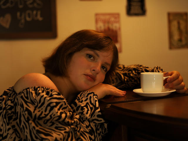 StaceyLex, a young woman with short brown hair, leans on a wooden table with a white cup and saucer in front of her. She wears a black-and-white zebra pattern off-shoulder top. Her expression is neutral, with slightly parted lips and half-closed eyes. The warm, yellowish light casts soft shadows. The table's dark wood contrasts with her top and the white cup. The background includes blurred, framed art and a chalkboard with text. Her right hand rests on the table, while her left arm supports her head. The overall composition is relaxed and intimate.